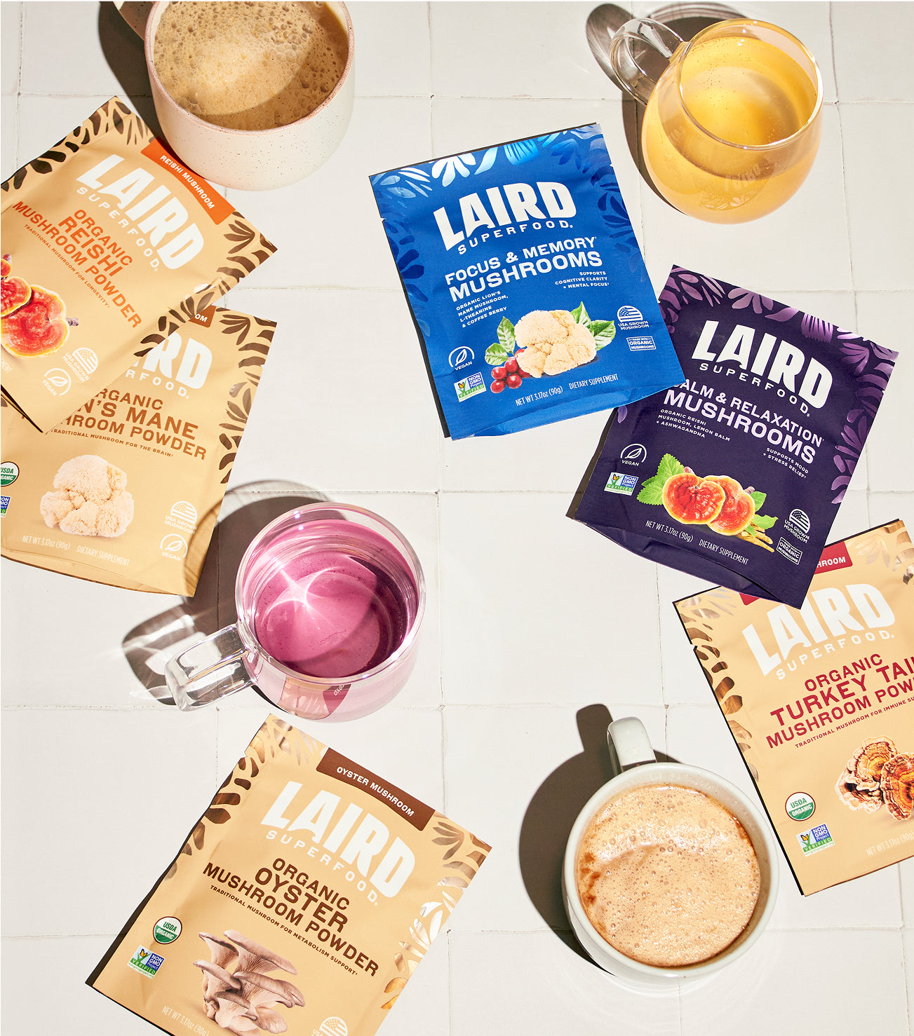 Collection of Laird Superfood mushroom powders with some made into drinks held in mugs on a white surface.