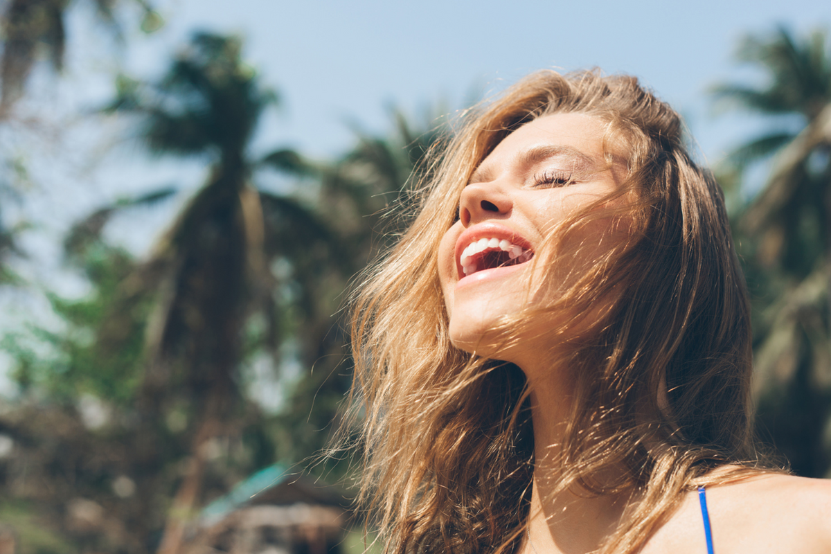 Woman with brown hair smiling with eyes closed in a tropical setting facing the sun