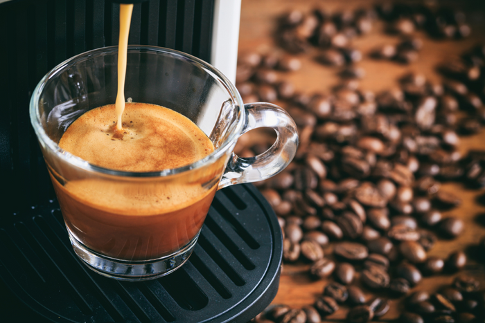 Espresso being brewed into a glass cup with coffee beans surrounding it