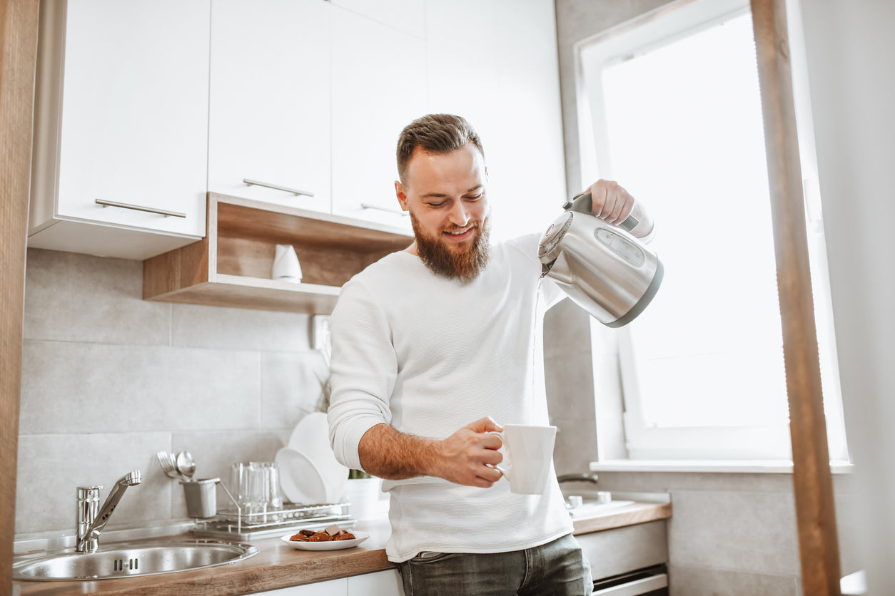 Man stands in kitchen pouring hot water into a mug to make an instant latte. 