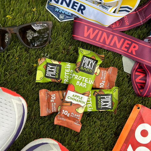 Apple Cinnawin Protein Bars on grass near winners medals, running shoes and sunglasses