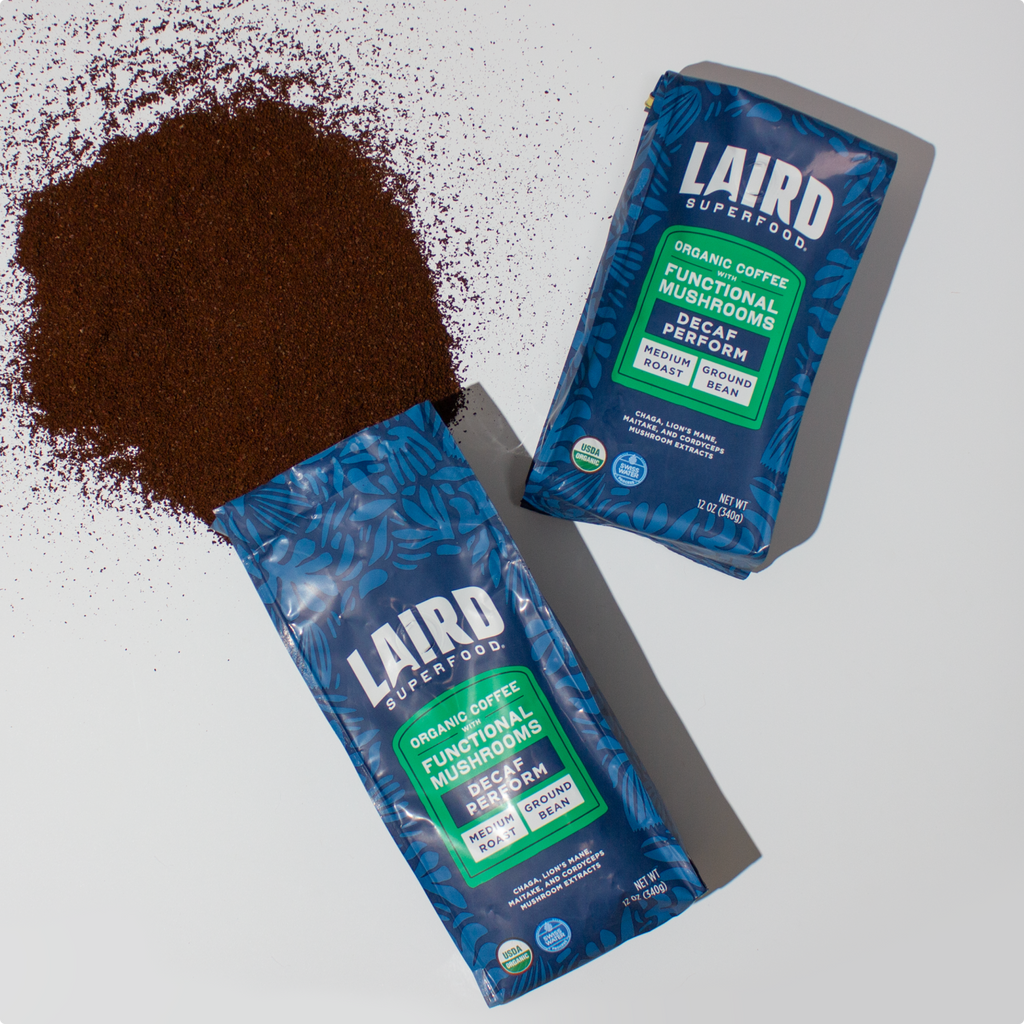 Two blue bags of Laird Superfood organic coffee with functional mushrooms, one spilled medium roast ground coffee on a white surface.