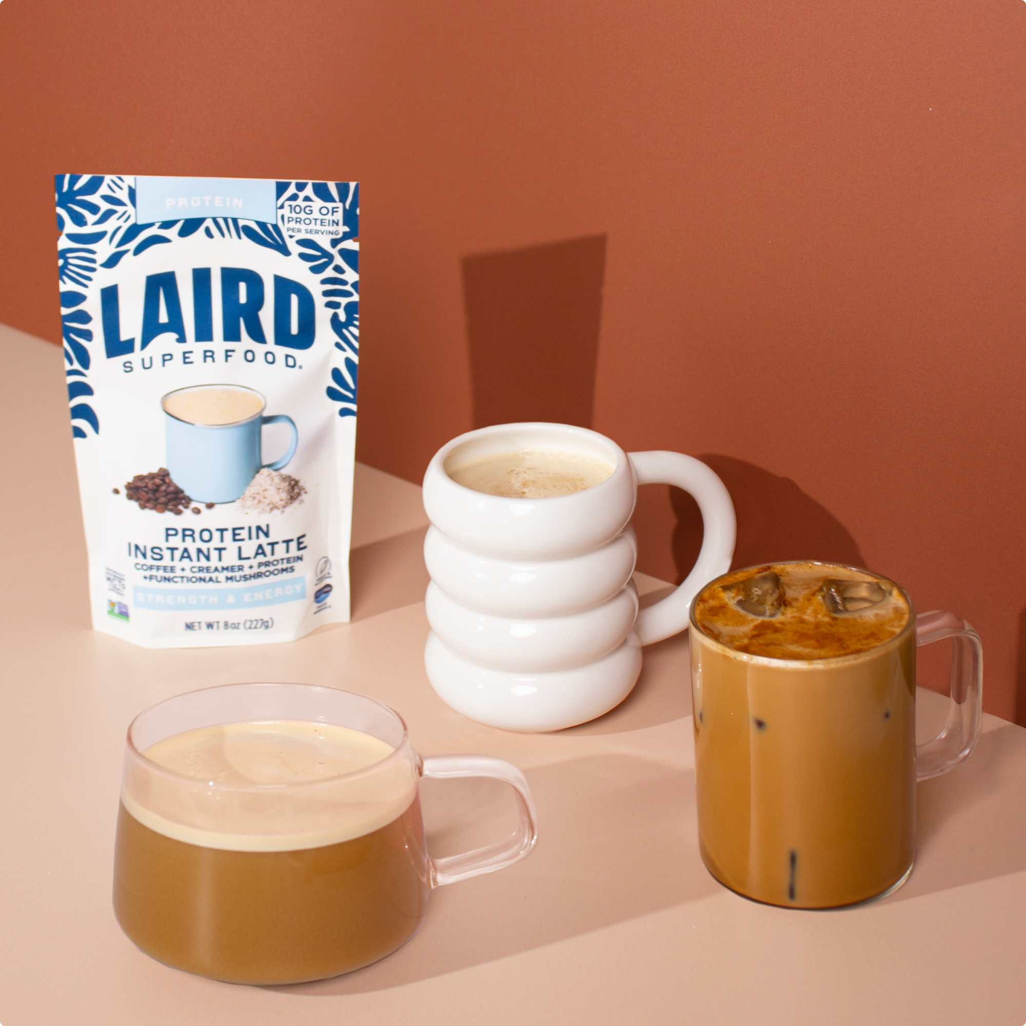 Laird Superfood protein latte bag displayed next to two clear mugs one with an ice latte and a white mug containing the instant latte