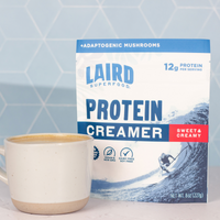 Laird Superfood Protein and Creamer mixed with coffee in a frothy mug next to the packaging on a table